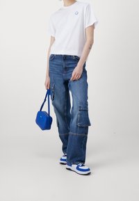 Hvid bomulds T-shirt med blåt logo, blå tværskuldertaske, løstsiddende blå denim cargo bukser, blå og hvide sneakers med polstret sål.