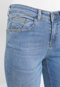 Jeans de denim azul claro con corte slim, que cuentan con un diseño clásico de cinco bolsillos y costuras en contraste. El desgaste visible añade textura.