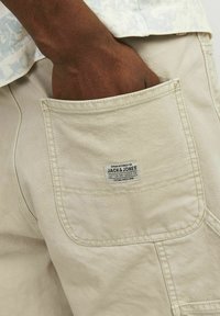 Tmavá ruka v zadní kapse béžových bavlněných kalhot s viditelnou etiketou Jack & Jones na kapse.