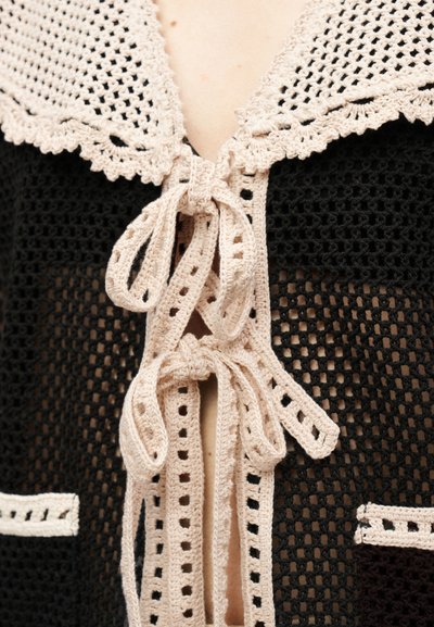 Μαύρο πλεκτό ένδυμα με μπεζ πλεκτό γιακά και κορδόνια δεμένα σε δύο φιόγκους στο στήθος ενός ατόμου.