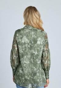 Camicia verde chiaro con motivi floreali, maniche trasparenti e collo a camicia. Presenta maniche a sbuffo arricciate e orlo dritto.