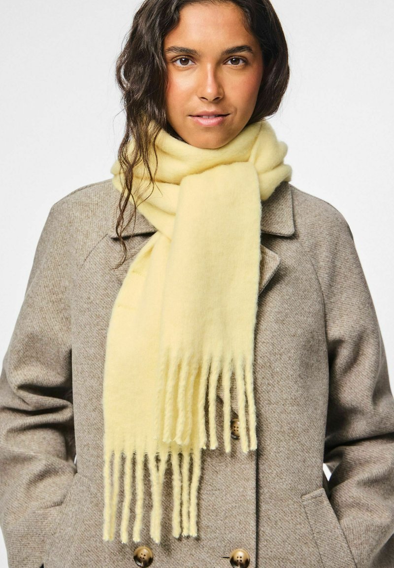 Donna con capelli scuri e mossi che indossa un cappotto di lana beige e una sciarpa frangiata di un giallo pallido avvolta attorno al collo.