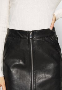 Jupe en cuir noir avec fermeture éclair frontale, présentant une texture lisse et un léger éclat. La ceinture est structurée et épouse étroitement la taille.