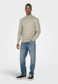 Hellgrauer Rollkragenpullover, lässige blaue Jeans mit Used-Effekten und schwarze Sneakers mit dicker weißer Sohle, stehend vor einem schlichten Hintergrund.