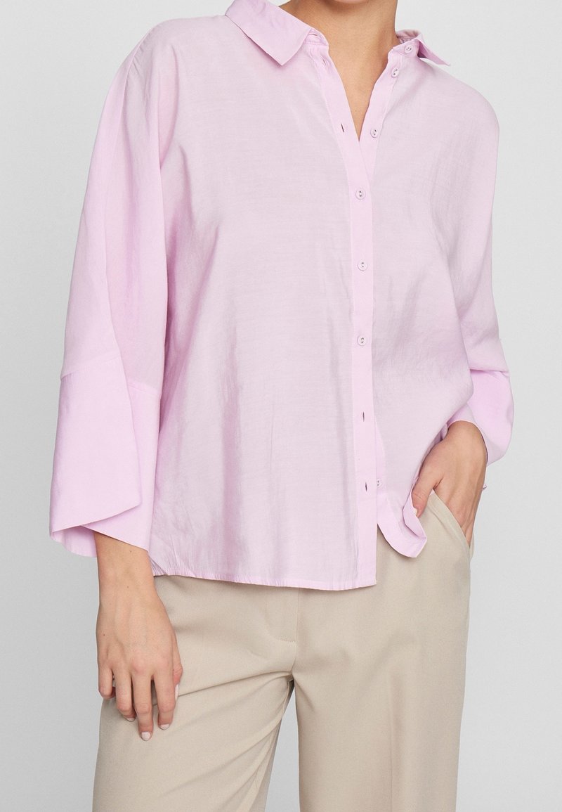 Femme portant une chemise ample rose clair à boutons avec des manches trois quarts et un pantalon beige, une main dans la poche, debout devant un fond uni.