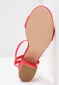 Sandale à talon haut en suède rouge avec un bout arrondi, une fine bride de cheville et une semelle beige lisse présentant une texture subtile.