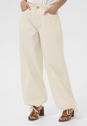 Pantalons larges beiges avec une taille à cordon, poches avant et détails de nouage aux chevilles, associés à des sandales à talons blocs marron.