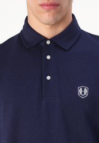 Tamsiai mėlynas polo marškinėliai, pagaminti iš tekstūruotos medžiagos. Yra klasikinis apykaklė, trys mygtukai ir siuvinėtas logotipas ant krūtinės.