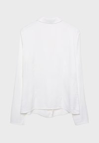 Blouse blanche à manches longues avec col, couture dorsale visible et ourlet légèrement incurvé, présentée sur un fond gris clair uni.