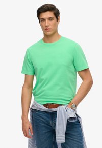 Mintgrünes Baumwoll-T-Shirt mit einem Rundhalsausschnitt, kurzen Ärmeln, kombiniert mit blauen Jeans und einem hellblau gestreiften Hemd, das an der Taille gebunden ist.