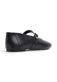 Chaussure plate en cuir noir avec un bout pointu, une lanière à la cheville et un détail de boucle ronde. Présente un petit talon empilé et une texture lisse.