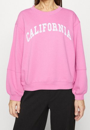 Person trägt ein pinkfarbenes Sweatshirt mit dem Schriftzug "CALIFORNIA" in weißen Buchstaben auf der Brust und eine schwarze Hose, steht mit entspannten Armen.