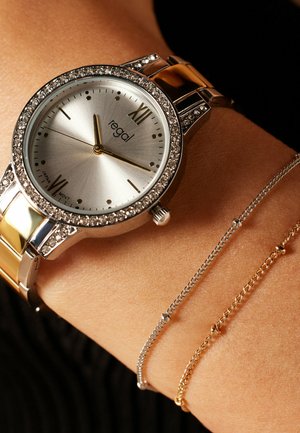 Silver watch with stone-encrusted bezel, Roman numerals, and metallic band. Features thin silver and gold chain bracelets beside it.