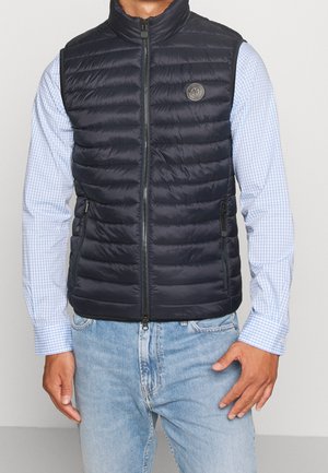 Mężczyzna ubrany w granatową pikowaną kamizelkę bez rękawów na jasnoniebieskiej koszuli w kratę i jasnych dżinsach, stojący z jedną ręką w kieszeni.