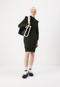 Robe tricotée vert olive avec une silhouette ajustée, ornée de volants sur les épaules. Le modèle porte un sac fourre-tout noir avec une bordure en fausse fourrure crème.