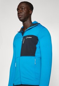 Kék cipzáras dzseki texturált anyagból és fekete részletekkel. Jellemzője a mellkasi cipzáras zseb és a állítható kapucni. A logó a mellkason látható.