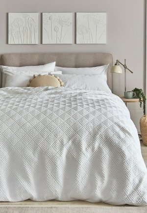 Bett mit weißer, strukturierter Bettwäsche, beigem Kopfteil, dekorativem rundem Kissen, Nachttisch mit Lampe, Tasse und Pflanze sowie floralem Wandbild darüber.