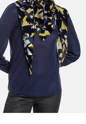 Persoon met een donkerblauw shirt met lange mouwen, zwarte broek en een multicolor sjaal met abstract patroon om de hals.