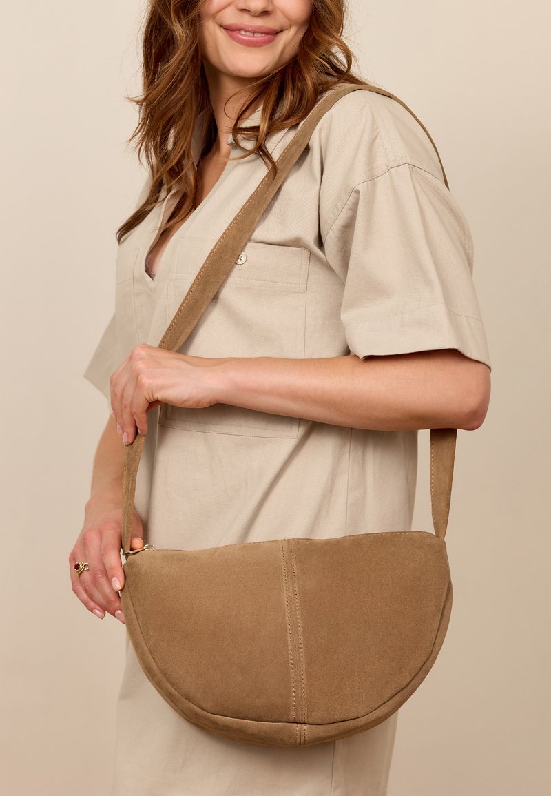 Ruskea mokkanahkainen olkalaukku, puolikuun muotoinen, näkyvällä ompeleella ja pitkällä hihnalla, pukeutuneena neutraalin sävyiseen asuun.