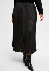 Svart plisserad midikjol i glänsande material, med elastisk midjeband och en slät textur, tillsammans med svarta ankelskor.