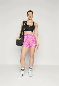 Μαύρο αθλητικό σουτιέν με λευκές ρίγες, ροζ σόρτς tie-dye, λευκές αθλητικές κάλτσες με μαύρες ρίγες, κρατώντας μια μαύρη τσάντα.