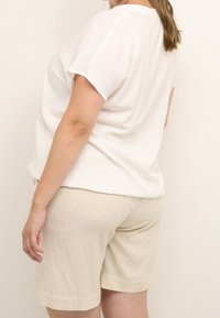 Haut blanc à manches courtes avec une finition texturée, associé à un short beige clair à mi-cuisses. Les deux vêtements sont amples et décontractés.