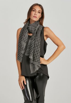 Vrouw met een mouwloos zwart topje, glanzende zwarte broek en een zwart-witte sjaal met hondentandmotief rond haar nek.