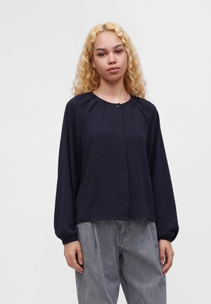 Jeune femme aux cheveux blonds bouclés portant un chemisier noir ample à manches longues et un pantalon gris sur un fond clair uni.