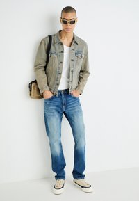 Ljust beige denimjacka med två framfickor, vit T-shirt, urblekta blåjeans och beige sneakers med marinblå accenter samt svarta solglasögon.
