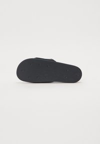 Musta kumisaappaan pohja, jossa on teksturoitu pinta, pyöreitä pitoäppiä ja kohotettu kuvio. Minimalistinen muotoilu ilman lisäkoristeita.