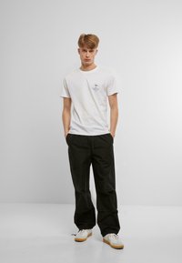 T-shirt en coton blanc avec un petit motif noir sur la poitrine gauche et un pantalon large noir. Le mannequin porte des baskets blanches avec des semelles orange.