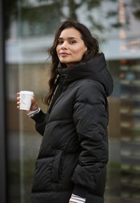 Schwarze Daunenjacke mit hohem Kragen, verstellbarem Kapuzenzug und Fronttaschen. Das Model hält eine Kaffeetasse und trägt darauf gestreifte Ärmel.