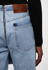 Jeans en denim bleu clair avec une fermeture éclair en métal, arbore un patch en cuir brun étiqueté "KSENIA SCHNAIDER" et une poche arrière avec une petite tache.