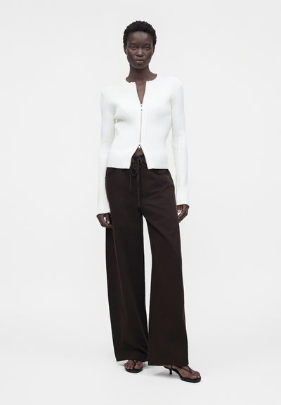 Γυναίκα στέκεται φορώντας ένα λευκό ριπ πλεκτό πουλόβερ με φερμουάρ, φαρδιά μαύρη παντελόνα με κορδόνι και μαύρα σανδάλια με τακούνι σε απλό φόντο.