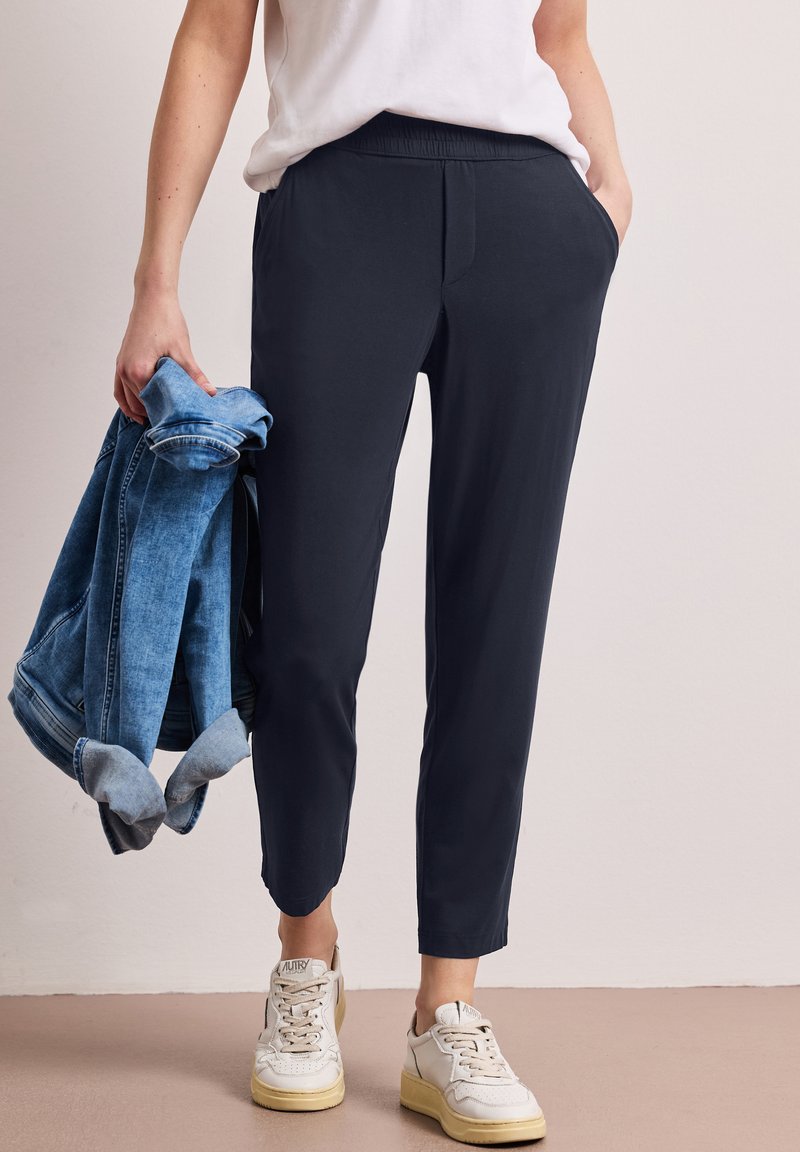 Person iført sorte bukser med elastisk talje og korte ben, hvide sneakers og en hvid skjorte, der holder en rullet denimjakke i den ene hånd.