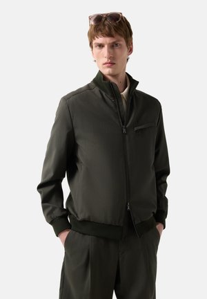 Ung mand iført mørkegrøn jakke med lynlås og matchende bukser, med solbriller hvilende på hans hoved, hænderne i lommerne, neutral baggrund.