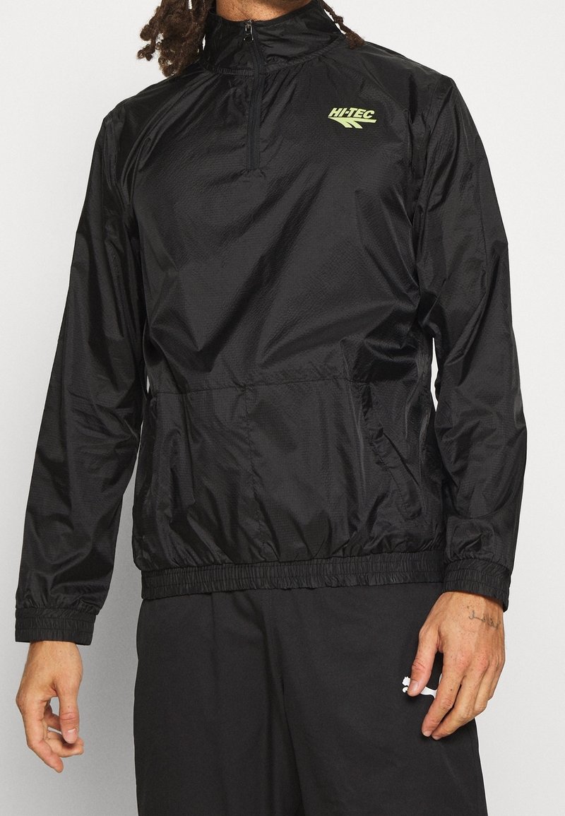 Homme portant un coupe-vent Hi-Tec noir à demi-fermeture éclair avec poignets et ourlet élastiques, associé à un pantalon noir, debout devant un fond uni.