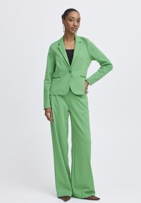 Vrouw staand met één hand op de heup, gekleed in een groen colbert en broek met wijde pijpen, een zwarte top, bruine puntige schoenen en hangende oorbellen.