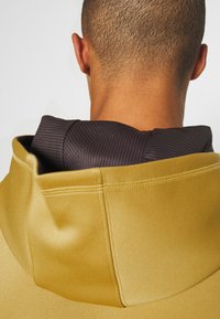 Huvtröja med en gul slät yta och en svart randig insida. Synliga sömdetaljer förhöjer designen.