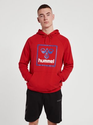 Młody mężczyzna w czerwonej bluzie z kapturem z niebieskim logo i napisem „hummel”, z rękami w przedniej kieszeni, w czarnych szortach, stojący na jednolitym tle.