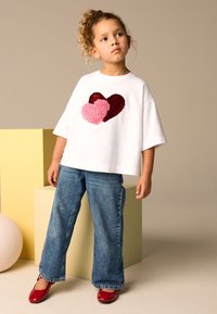 White short-sleeve t-shirt featuring a red and pink heart design, paired with blue denim flared jeans and red shoes. Set against neutral background.