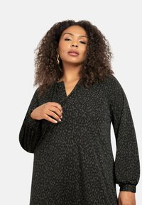 Šaty s dlouhým rukávem v tmavě zelené barvě s jemným abstraktním potiskem. Mají V-výstřih a elastické manžety pro uvolněný střih.