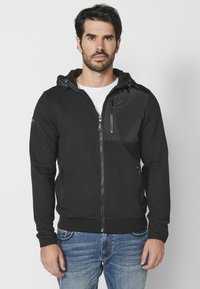 Felpa nera con zip, corpo testurizzato, pannello liscio sul petto, cappuccio regolabile e polsini aderenti; abbinata a jeans azzurri chiari.