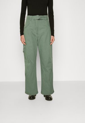 Calças cargo verdes com um design de perna larga, cinto de tecido, múltiplos bolsos e uma textura suave. Usadas com uma blusa preta de mangas longas e botas pretas.
