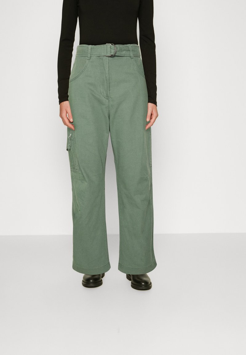 Calças cargo verdes com um design de perna larga, cinto de tecido, múltiplos bolsos e uma textura suave. Usadas com uma blusa preta de mangas longas e botas pretas.