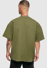 Olivově zelené oversized tričko s krátkým rukávem, žebrovaným kulatým výstřihem a plochými švy. Materiál vypadá hladce a má jemnou texturu.