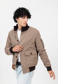 Mann passt seine braune Wolljacke mit Knöpfen und großer Vordertasche an, trägt ein weißes Hemd und hellblaue Jeans vor einem einfarbigen Hintergrund.