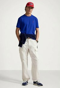 Νεαρός άνδρας που φοράει κόκκινο καπέλο, μπλε μπλουζάκι, λευκά cargo παντελόνια, ναυτικό πουλόβερ tied γύρω από τη μέση του και ναυτικά sneakers, στέκεται με τα χέρια στις τσέπες.