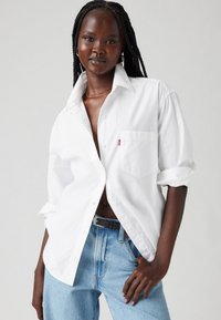 Chemise blanche à manches retroussées, poche poitrine unique, en coton doux, associée à un jean en denim bleu clair taille haute.
