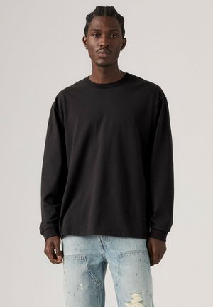 Mand med flettet hår iført en ensfarvet sort langærmet skjorte og lyseblå, revnede jeans, stående foran en ensfarvet lys baggrund.